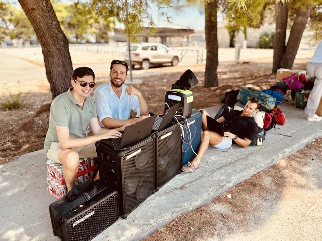 Friends working on laptops beside stacked travel cases outdoors.